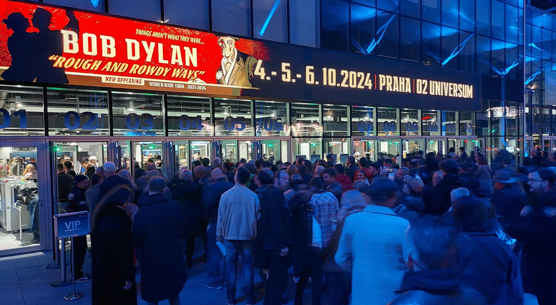 Velika množica ljudi pred vhodom v dvorano, nad vrati je reklama za koncert