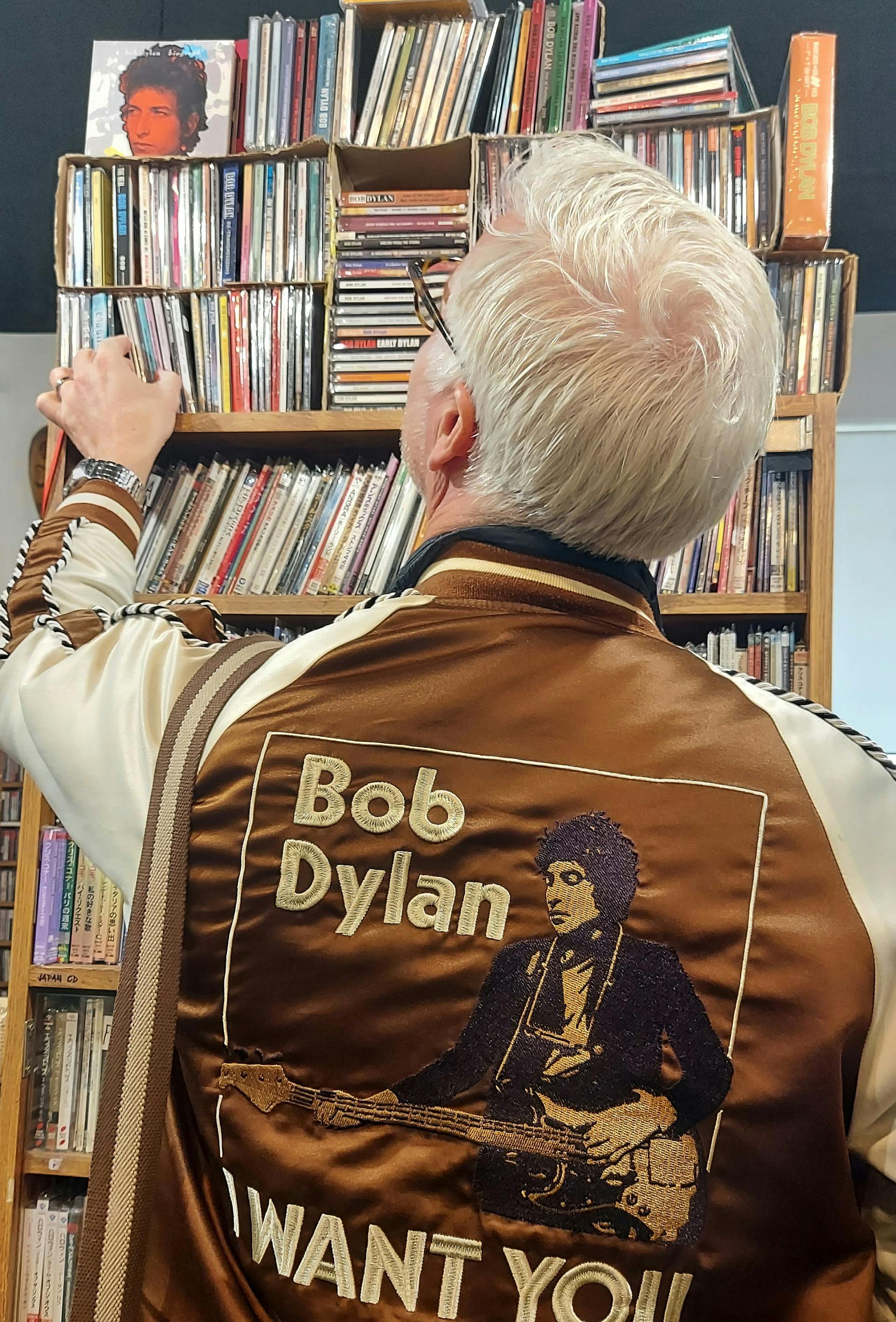 Moški, ki stoji pred stojalom s CD-ji, oblečeno ima jakno z Dylanovo podobo