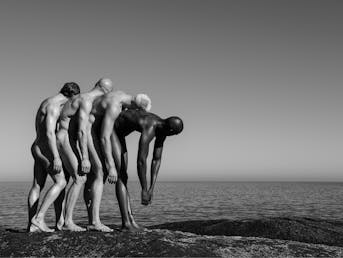 Črno-bela fotografija štirih golih moških na plaži, naslonjeni so drug na drugega in gledajo proti obzorju