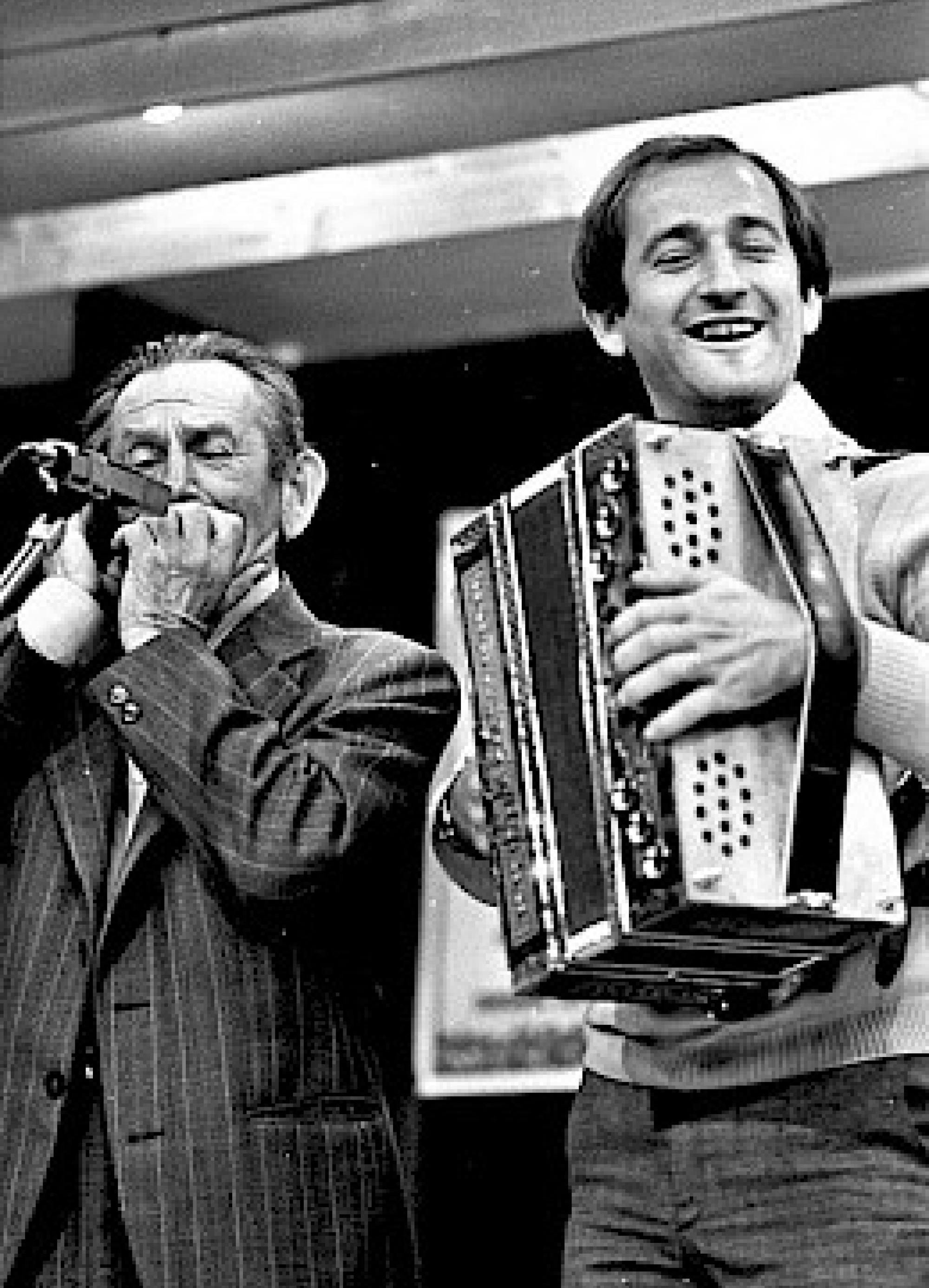 Moška stojita na odru, levi igra na orglice, desni na harmoniko.