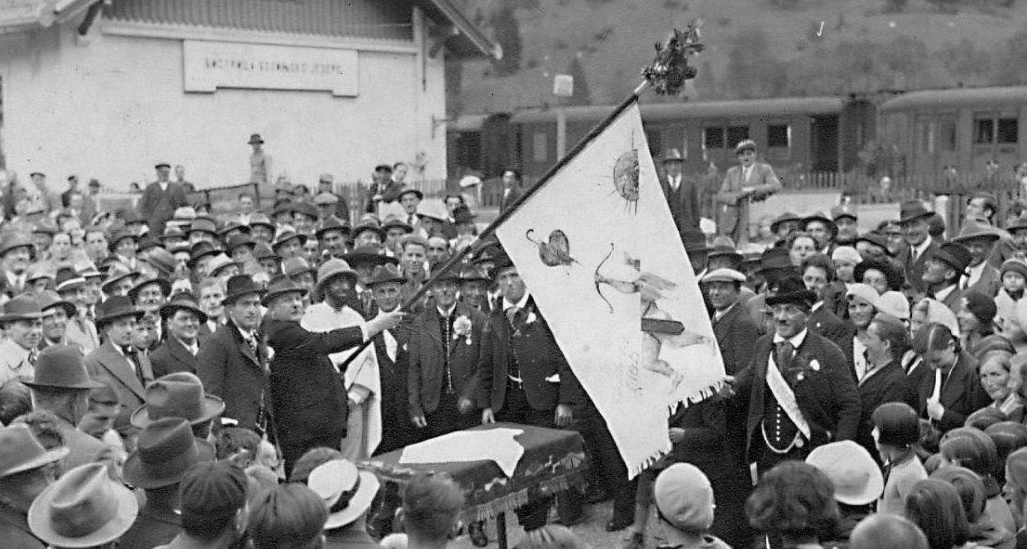 Gruča ljudi zbrana okrog mize nad katero držijo zastavo. Poleg zastavonoše stoji cesar v beli obleki, s klobukom.