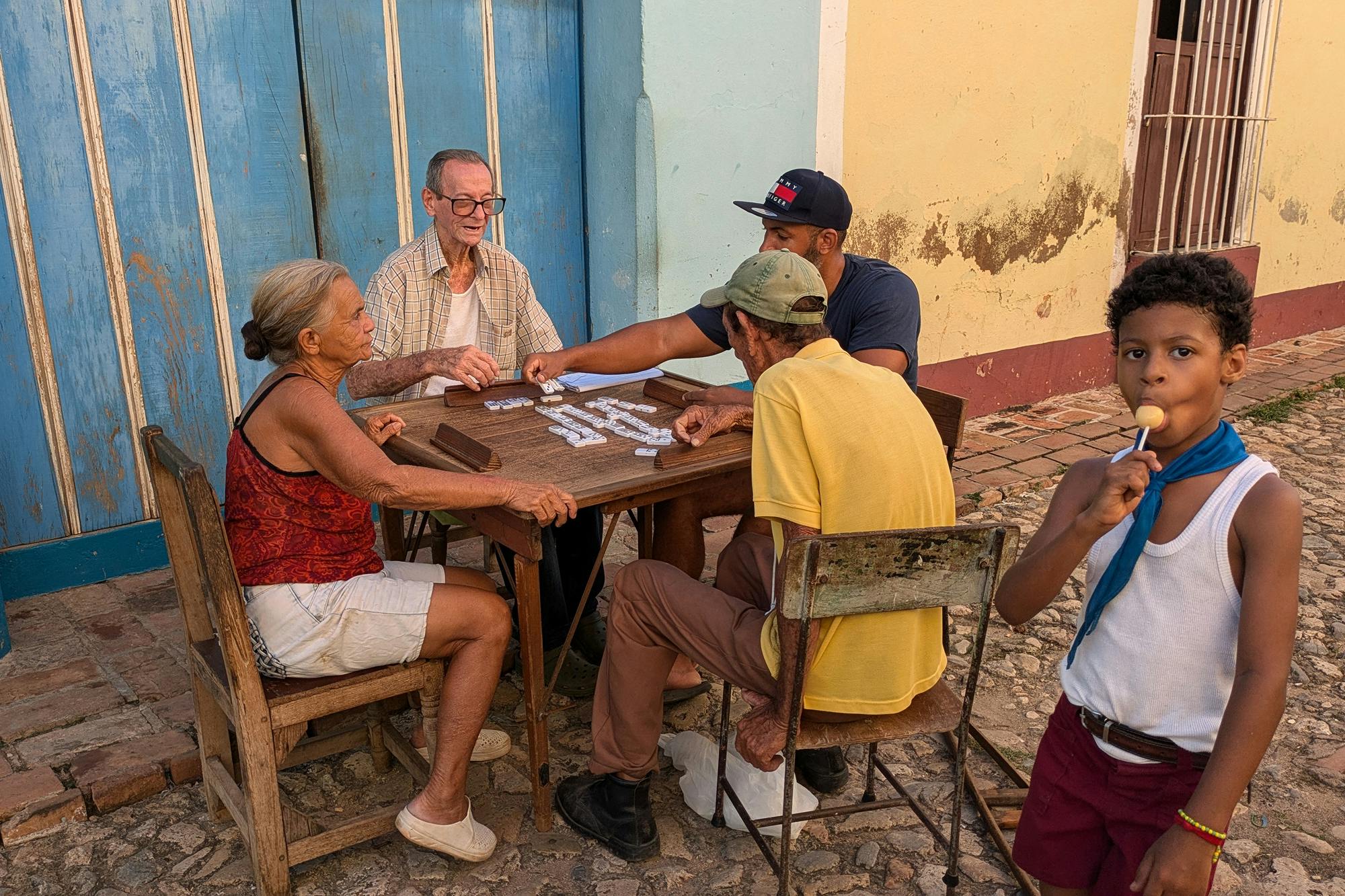 Štirje odrasli, ki sedijo za mizo na ulici in igrajo domino, pred njimi stoji majhen fant z liziko