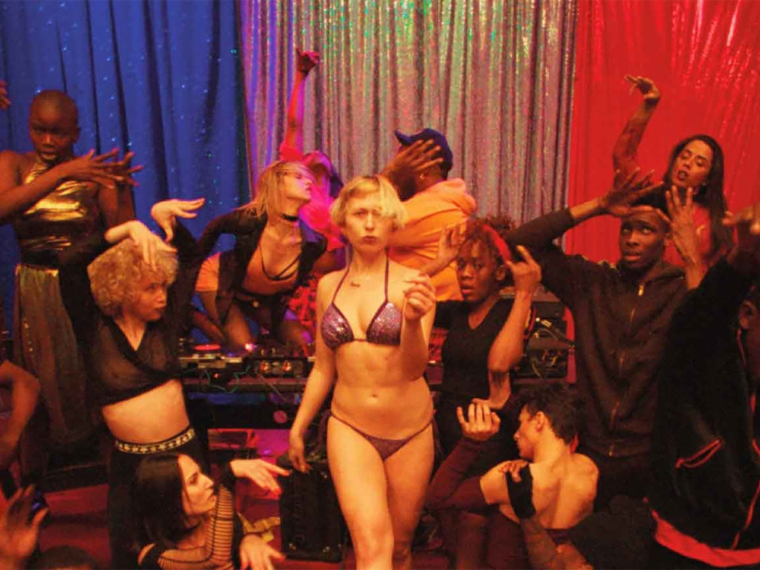 Plesalci na odru v razgibanem položaju. Na sredi ženska v kopalkah - gleda proti nam.