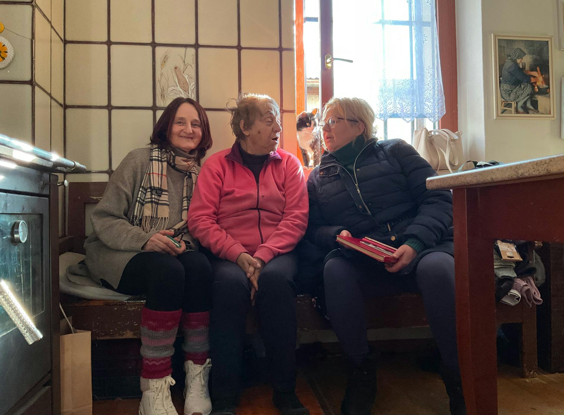 Silvana Paletti, Zvonka Simčič in Nina Kokelj sedijo skupaj na klopi, zadaj je priprto okno, pri katerem sedi mačka