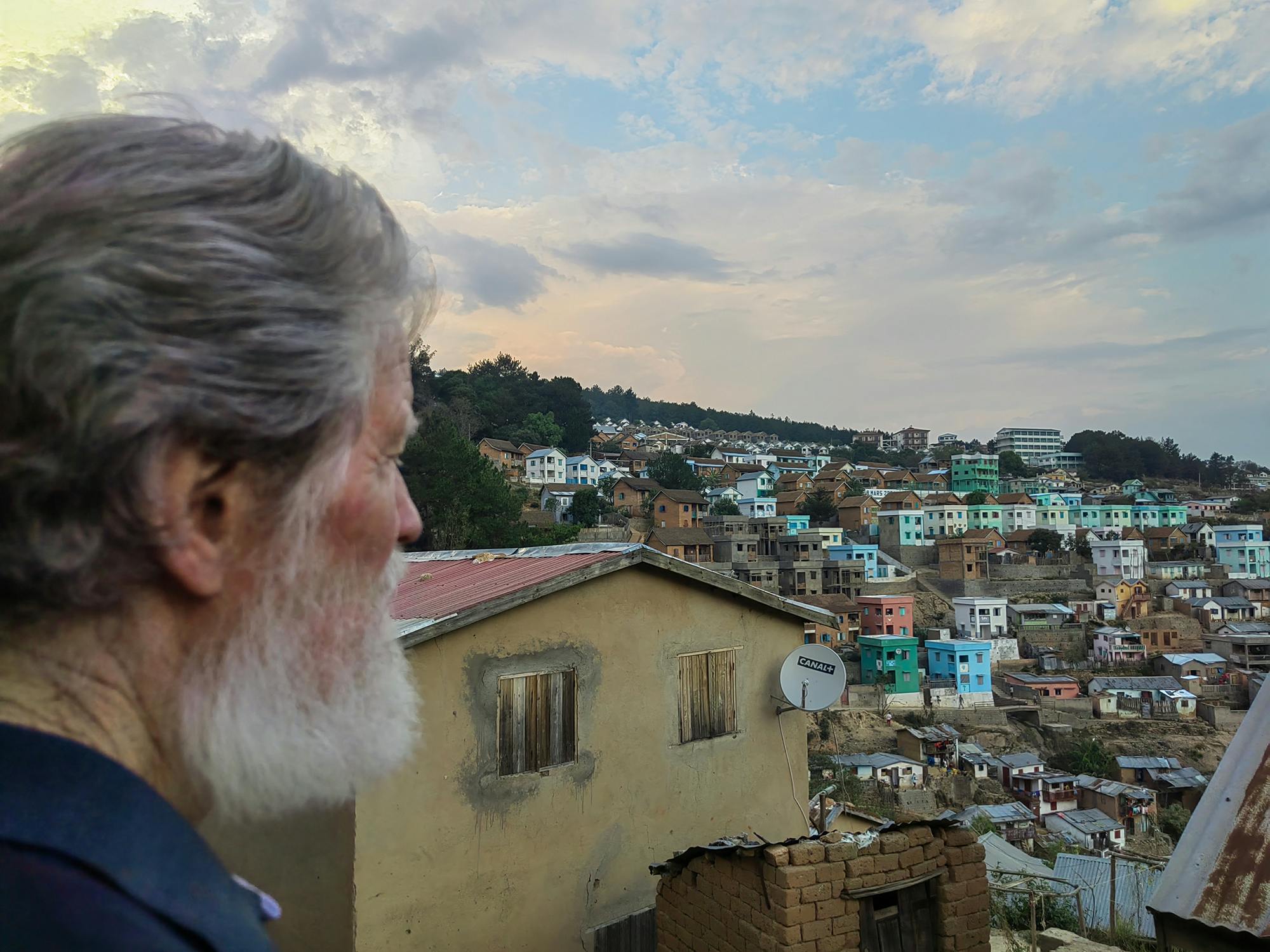 Pedro Opeka, fotografiran od strani, v ozadju pogled na barvite hiše mesta