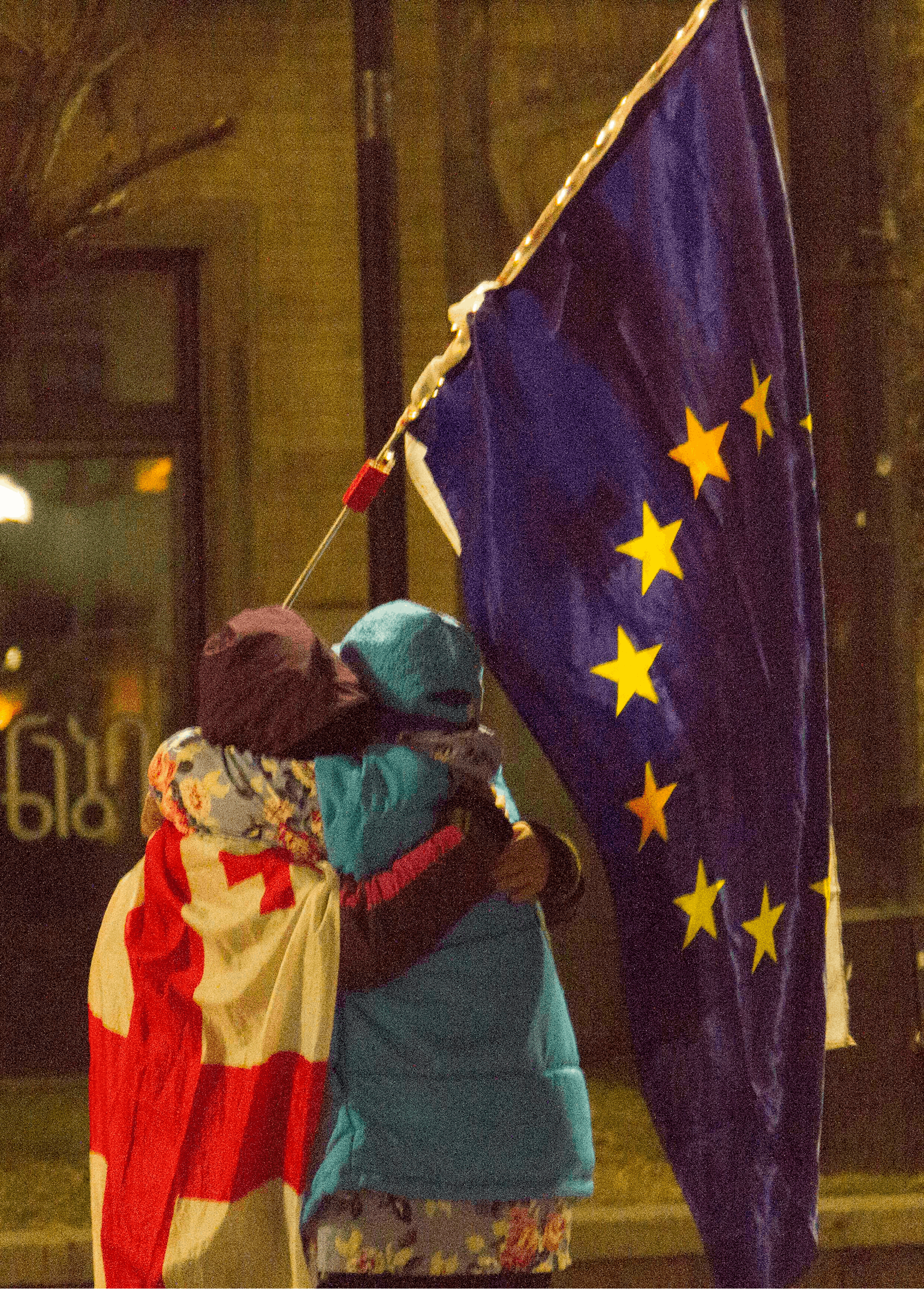 Dva protestnika, ki se objemata, eden ima okoli ramen ovito gruzijsko zastavo, drugi v rokah drži zastavo EU