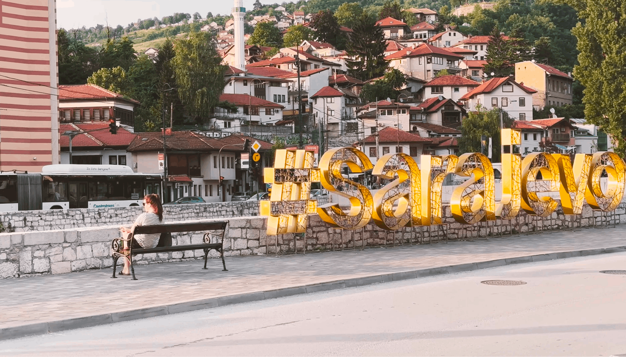 Ženska sedi na klopci zraven napisa #Sarajevo