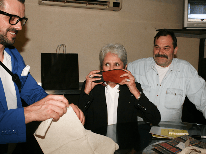 Joan Baez s čevljem pred obrazom, zraven nje sta dva moška