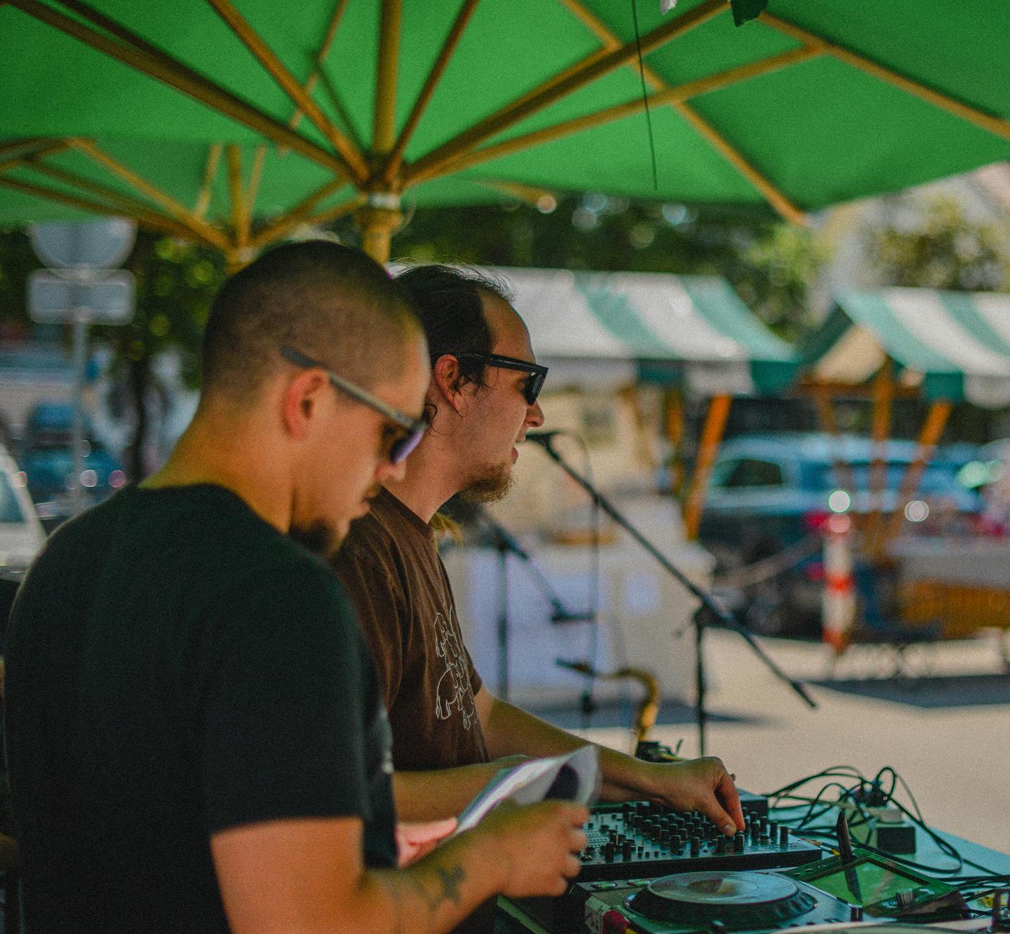 DJ vrti glasbo za mešalno mizo na stojnici pod zelenim senčnikom na tržnici Moste.
