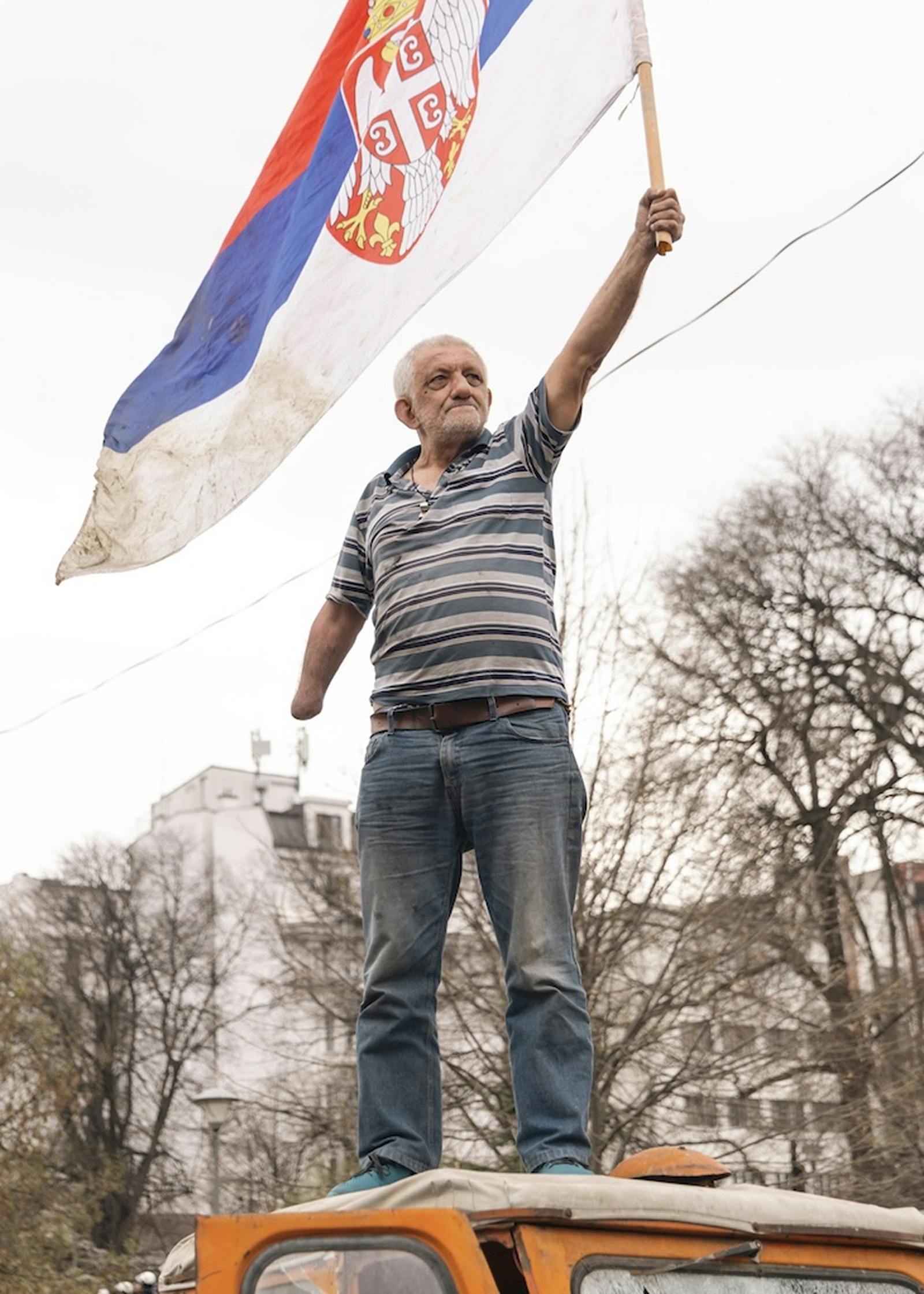 Starejši moški z amputirano roko na strehi avtomobila, ki maha s srbsko zastavo.