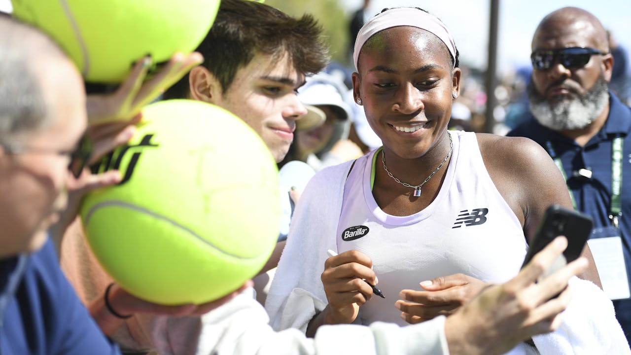 Coco Gauff takes a selfie with a fan and signs tennis balls.