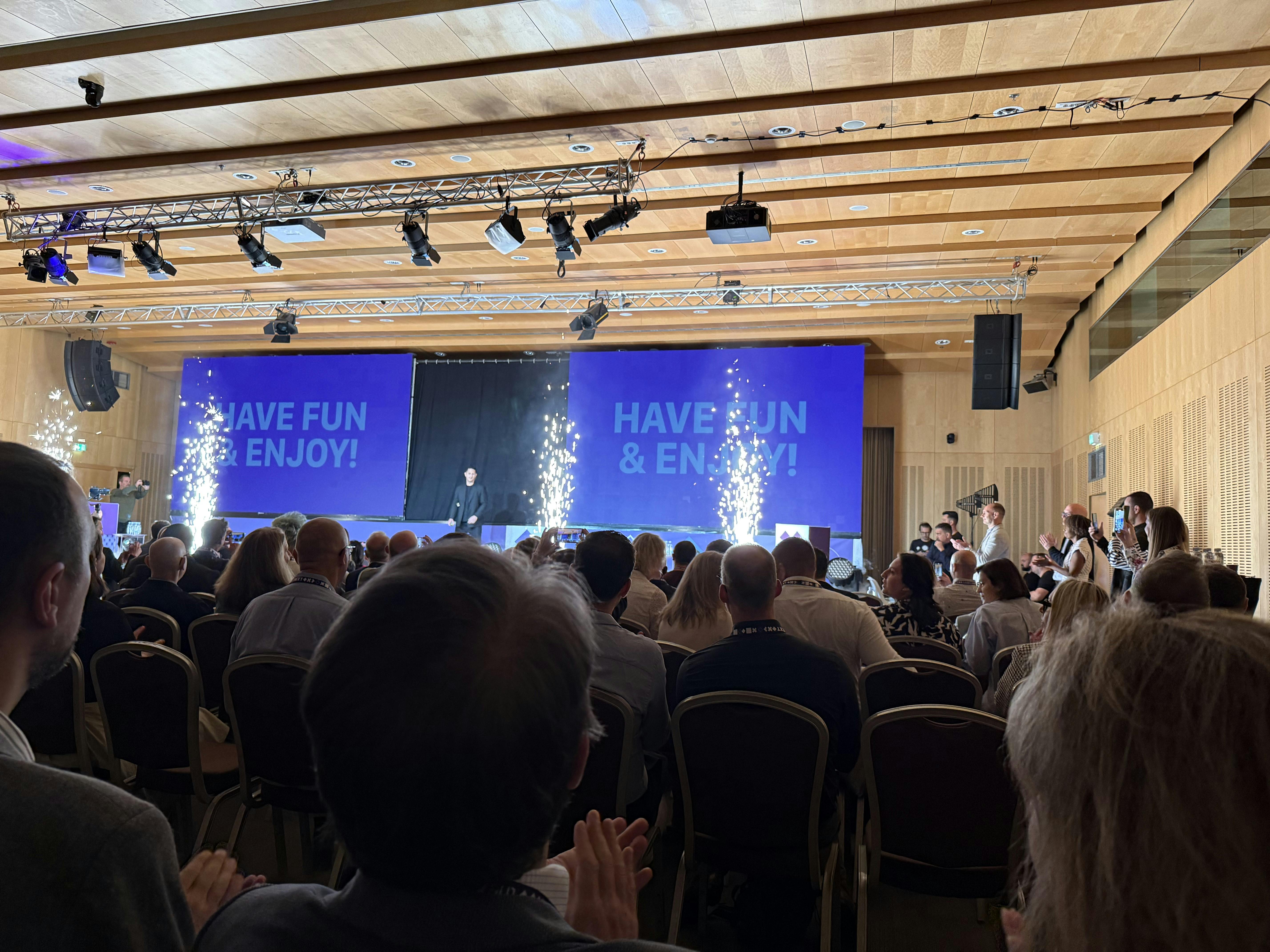 A large indoor conference room filled with seated attendees watching a stage presentation. On stage, a person stands in front of two large screens displaying the message "HAVE FUN & ENJOY!" Bright stage lights and sparkler-style pyrotechnics add excitement to the scene.