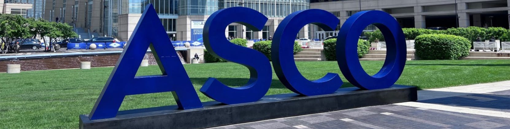 Large blue 3D letters spelling “ASCO” stand prominently in front of the glass-paneled McCormick Place convention center in Chicago, under a clear blue sky. The setting indicates the annual ASCO 2025 conference, with event signage visible in the background and attendees milling near the venue.