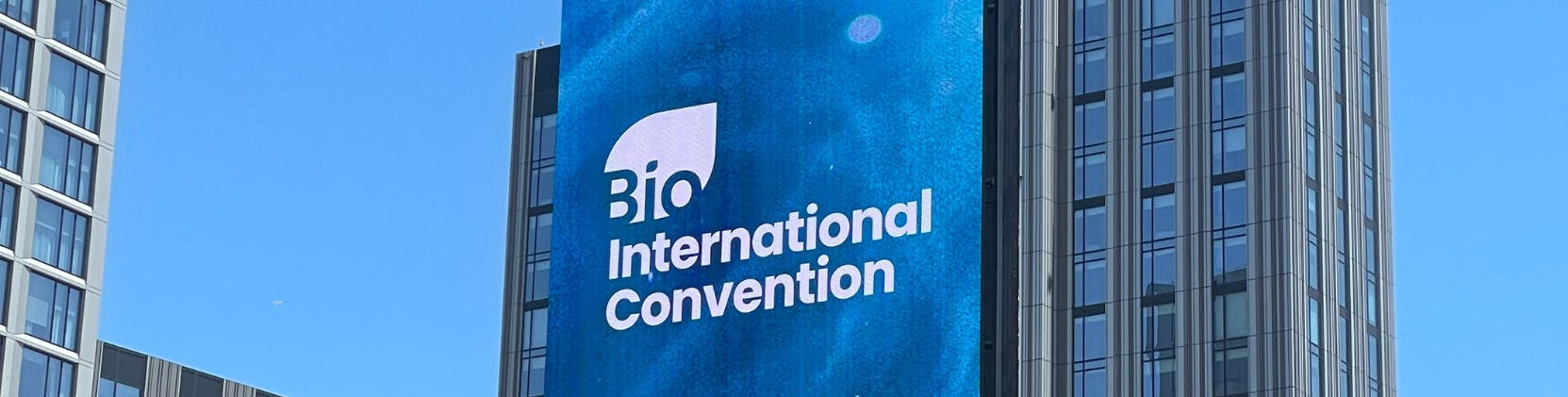 A tall digital billboard on the side of a modern high-rise building displays the logo and text “Bio International Convention” against a blue abstract background. The surrounding buildings have glass windows and vertical architectural lines, under a clear blue sky.
