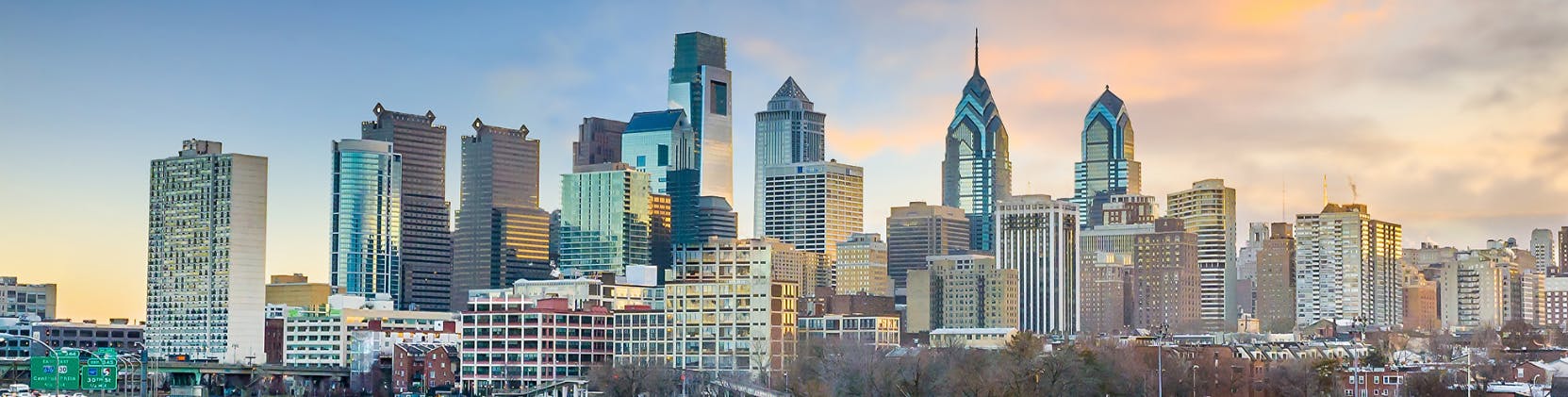 Panoramic view of downtown Philadelphia at sunset, showing modern skyscrapers and historic buildings reflecting warm evening light under a partly cloudy sky.