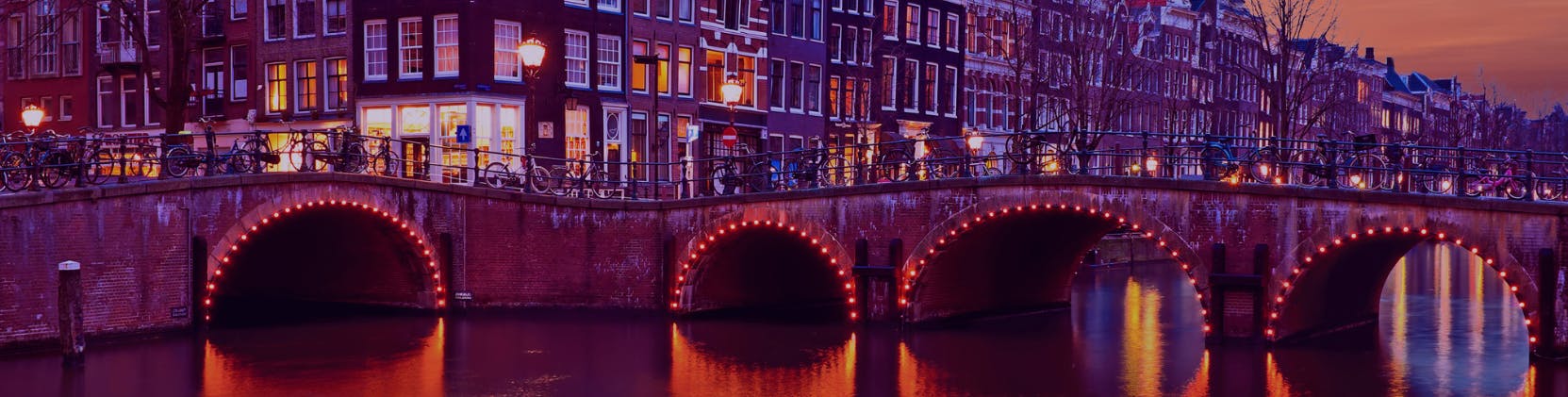 A picturesque evening view of a canal bridge in Amsterdam, illuminated with warm orange and red lights reflecting on the water, with bicycles lined up along the bridge and traditional Dutch townhouses in the background under a twilight sky.