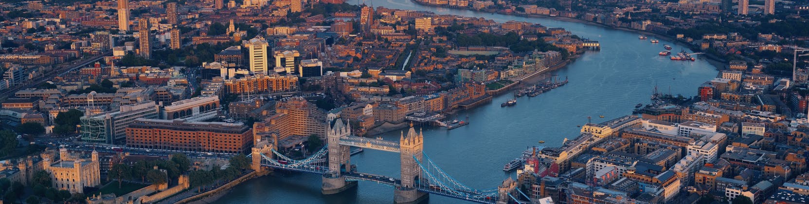 Aerial view of London at sunset featuring the iconic Tower Bridge spanning the River Thames. The cityscape is bathed in warm light, showcasing a blend of historic and modern architecture, including the Tower of London in the foreground and various buildings lining the riverbanks. Boats are visible on the water, and the dense urban layout highlights London’s vibrant infrastructure.