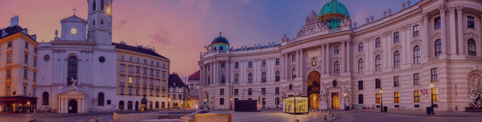 A panoramic view of Vienna’s historic Michaelerplatz at dusk, featuring the Baroque façade and green dome of the Hofburg Palace on the right, warmly lit buildings on the left, and a soft pink and purple evening sky overhead.