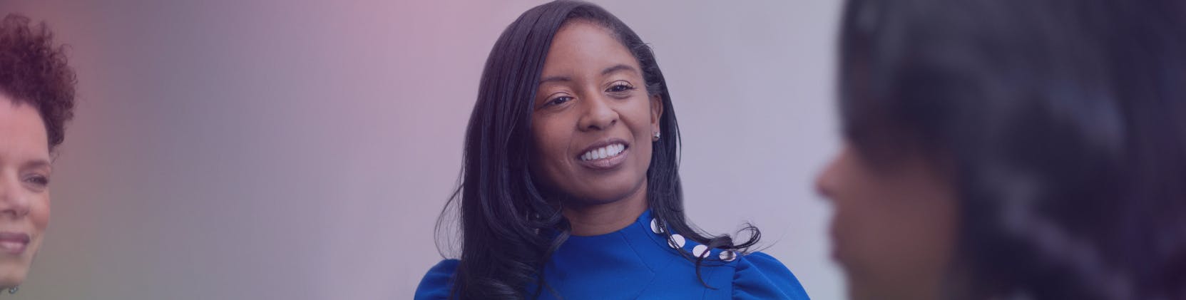 A woman in a vibrant blue dress, smiling and engaged in conversation with two others. The setting has a soft purple-toned filter, creating a warm and approachable atmosphere.