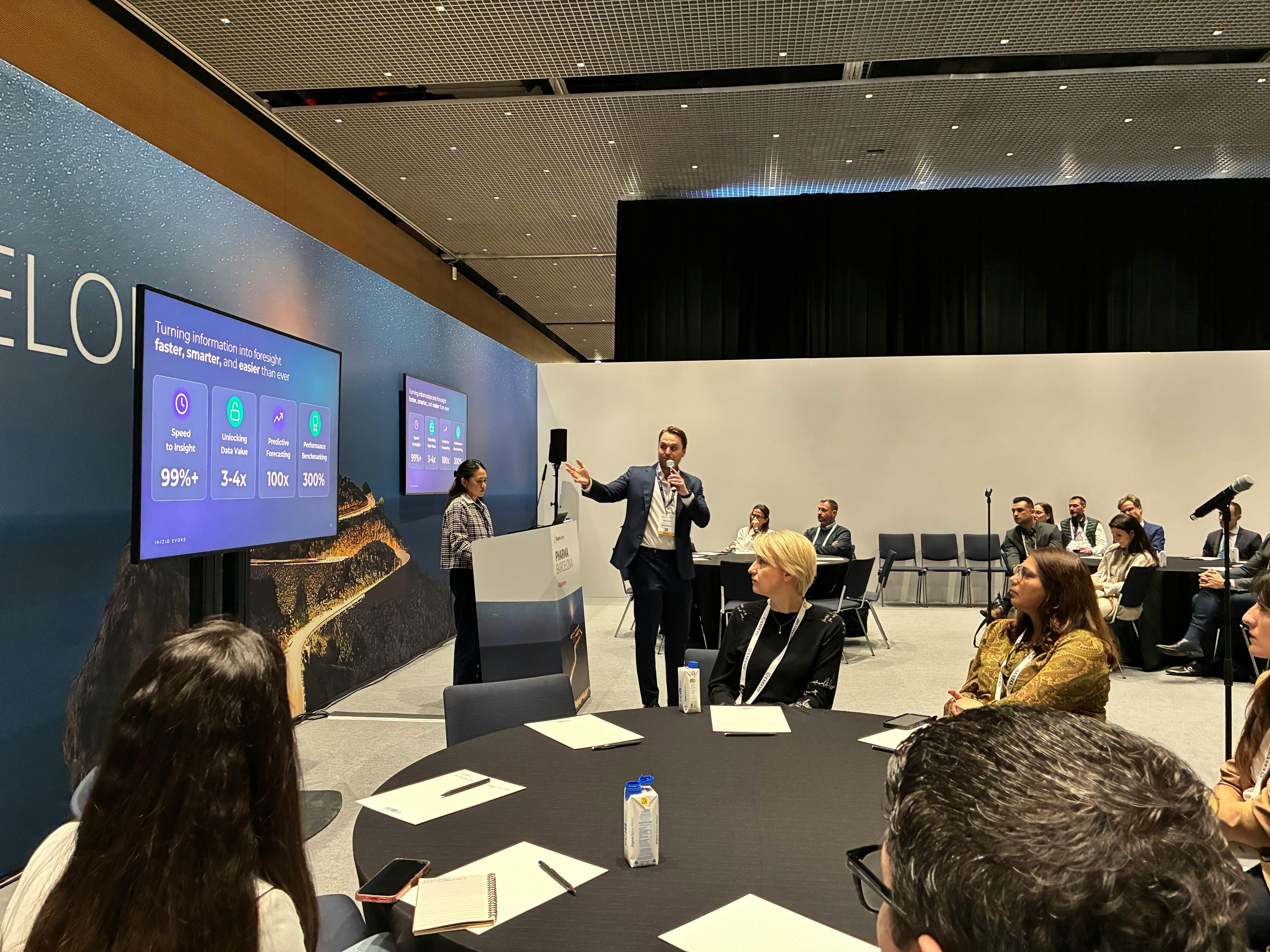 A conference session in a modern event space, where a man in a suit speaks into a microphone and gestures toward a seated audience at round tables.