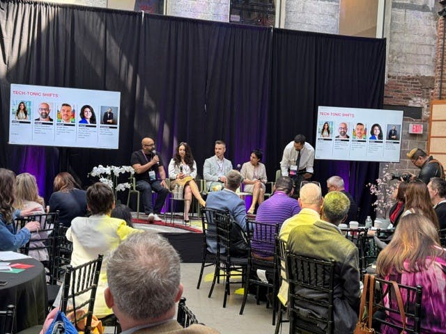 A panel discussion taking place on a small stage at a conference event, with five speakers seated in front of an audience. One speaker is holding a microphone while others listen. Two presentation screens on either side display the session title “Tech-Tonic Shifts” along with headshots and names of the panelists. Attendees sit at round tables facing the stage, and a photographer stands to the side capturing the event. The space has an industrial-style interior with exposed brick and draped curtains behind the speakers.