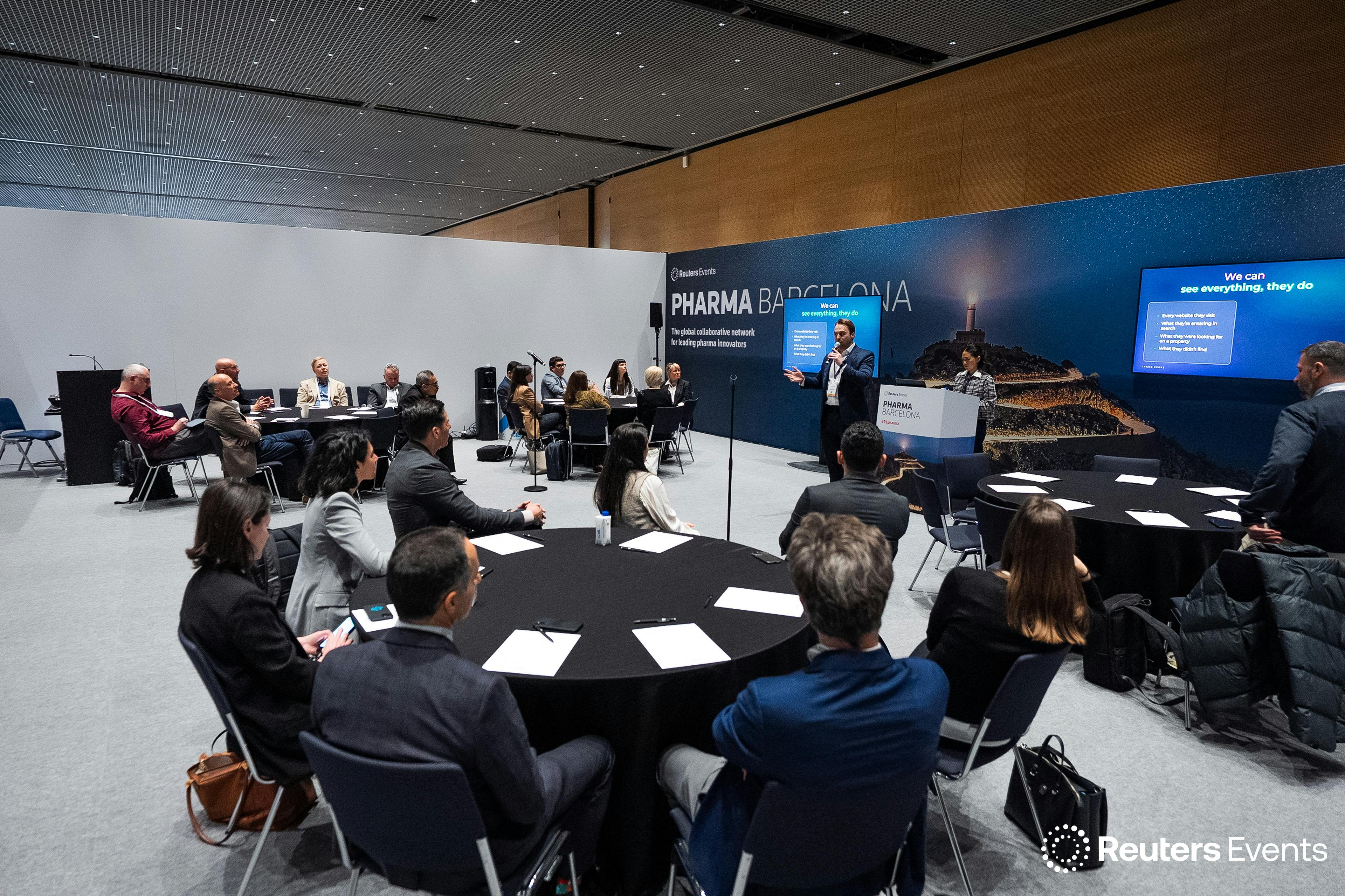 Audience members seated at round tables listen to a speaker presenting in an interactive session room at Reuters Events Pharma Barcelona. Two presentation screens display slides while attendees in business attire face the front of the room.