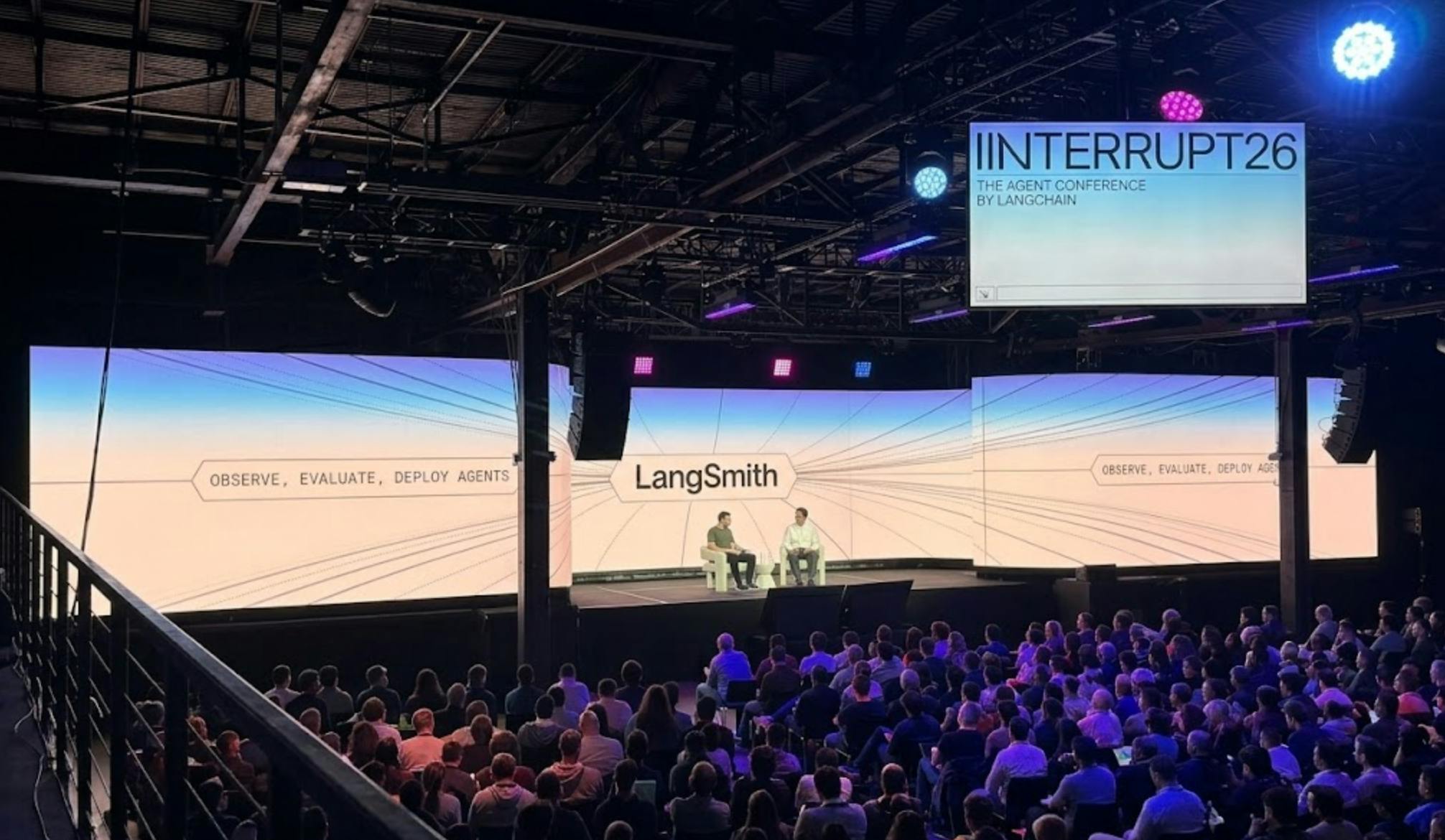 alt text for this image A large audience seated in a conference venue watches two speakers on stage beneath large screens displaying “LangSmith” and the slogan “Observe, Evaluate, Deploy Agents.” A hanging sign reads “INTERRUPT26 – The Agent Conference by LangChain,” with colorful stage lighting illuminating the industrial-style event space.