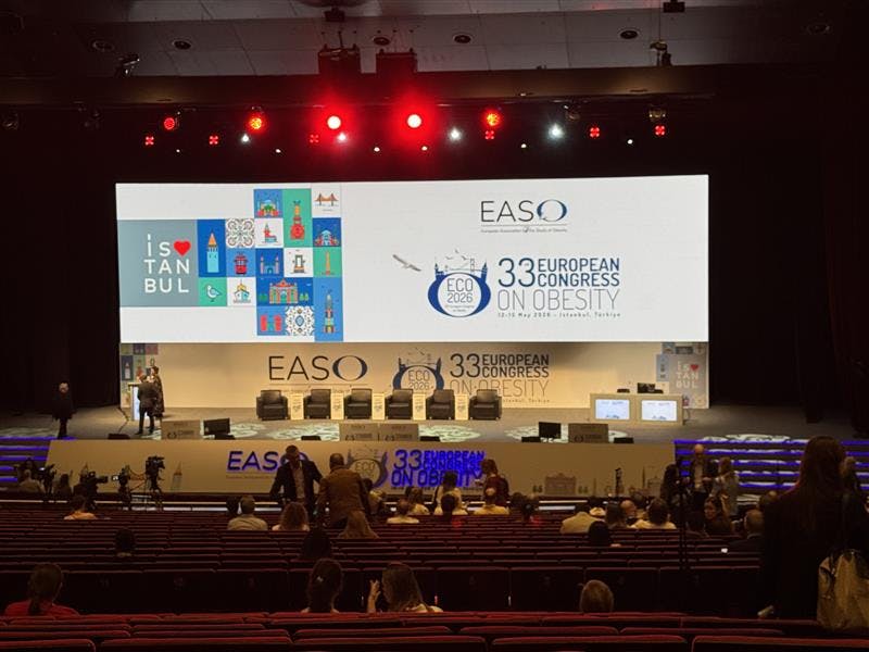 Wide view of a conference auditorium with attendees seated and gathering before a session. A large stage features branding for the European Association for the Study of Obesity (EASO) and the 33rd European Congress on Obesity (ECO 2026) in Istanbul, Turkey, displayed on a large screen above a panel discussion setup with several chairs. Red stage lighting illuminates the venue.