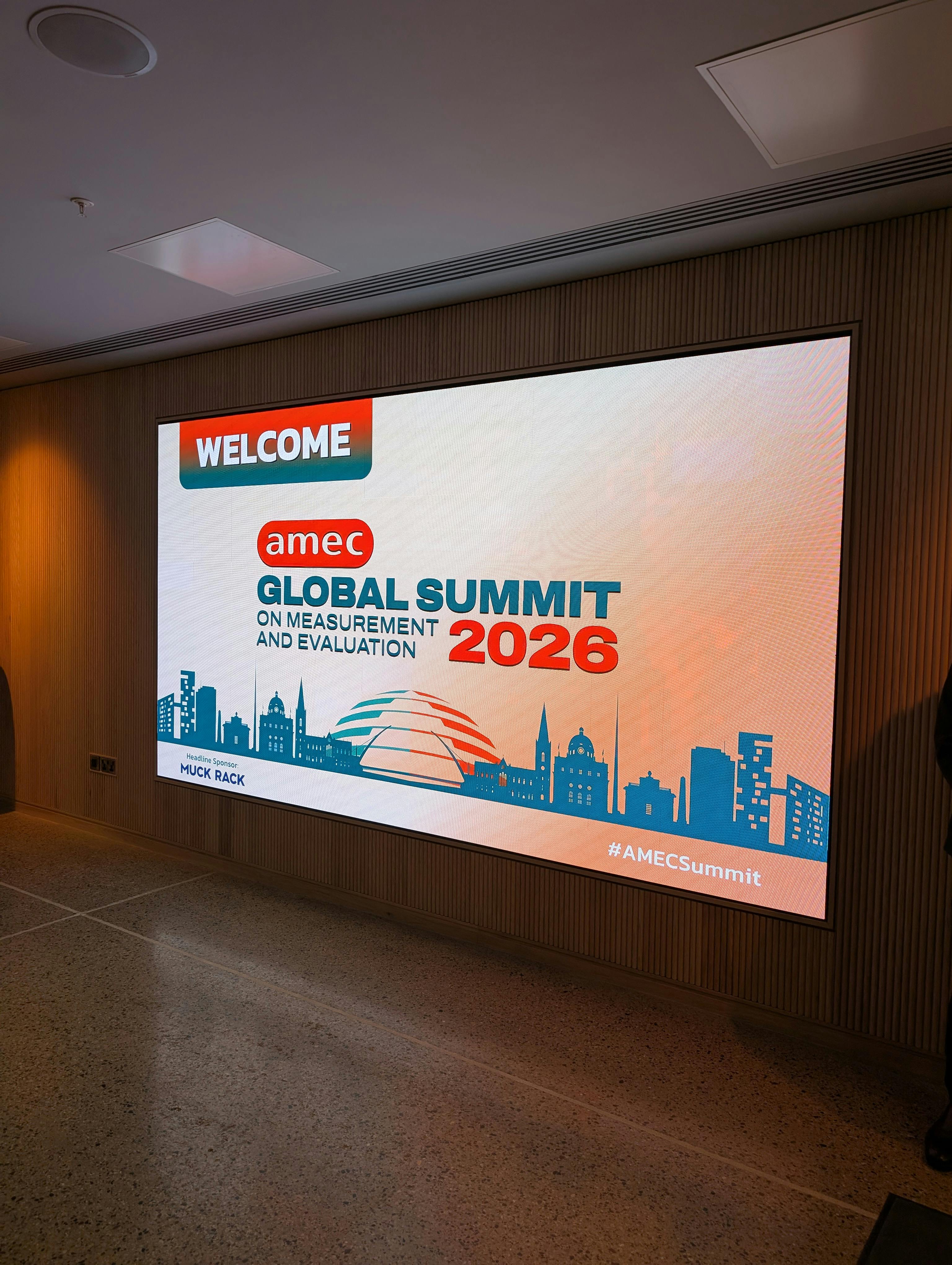 Photo of a large digital display welcoming attendees to the AMEC Global Summit on Measurement and Evaluation 2026, featuring a stylised city skyline and event branding.