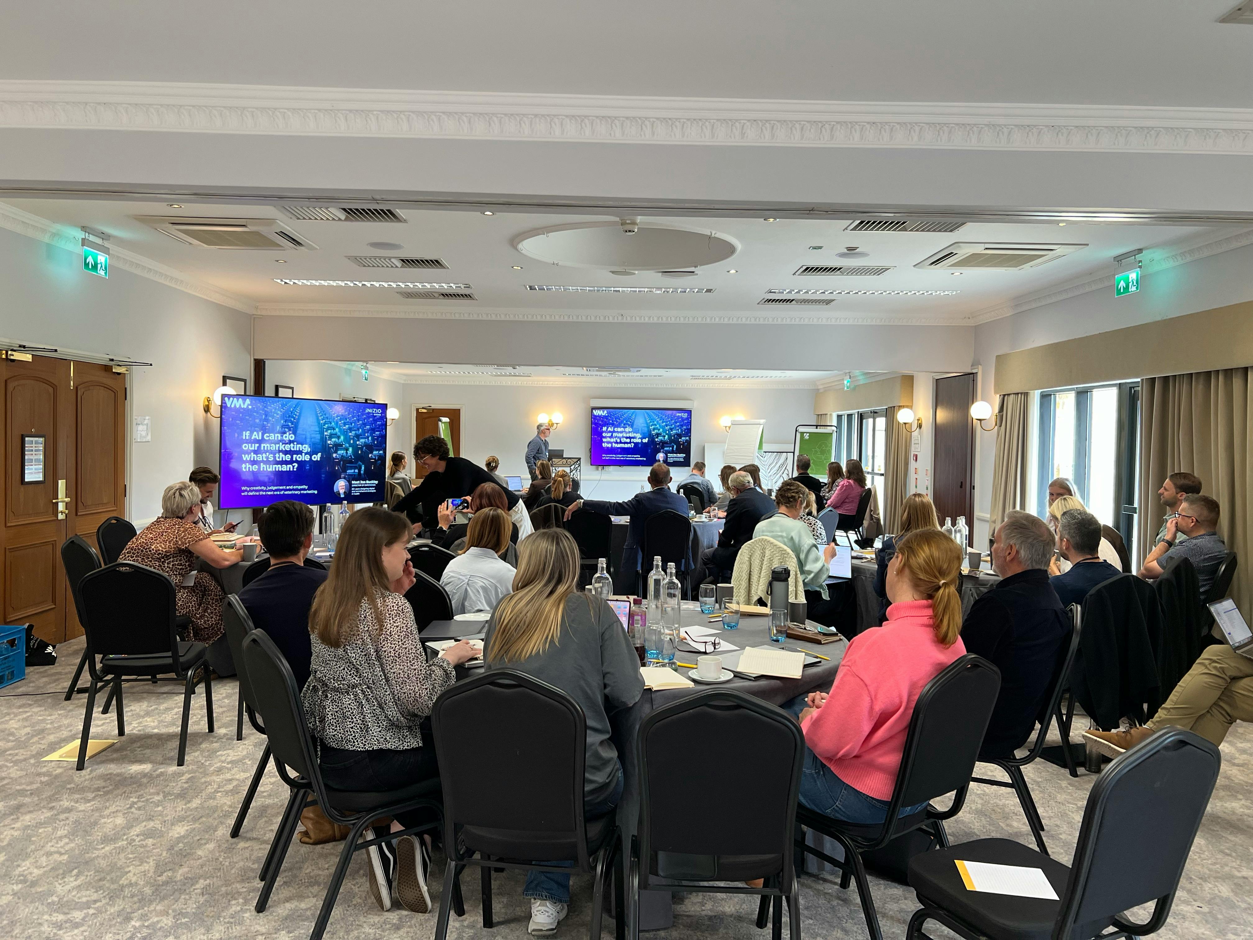 Attendees seated around tables at a Veterinary Marketing Association event, watching a presentation on AI and the role of humans in marketing.