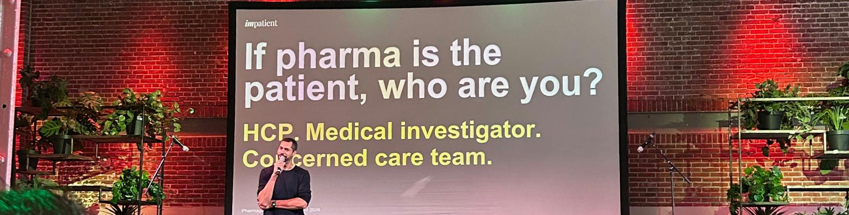 Speaker presenting on stage at Pharmageddon Europe, with a large screen displaying the question, “If pharma is the patient, who are you?” to an audience.