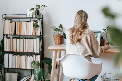 blonde woman typing at a desk with a bookshelf to her left and plants