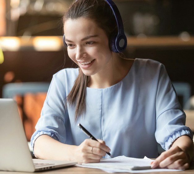 woman wearing headphones and a blue blouse looking at a computer screen and smiling while taking notes during a video conference