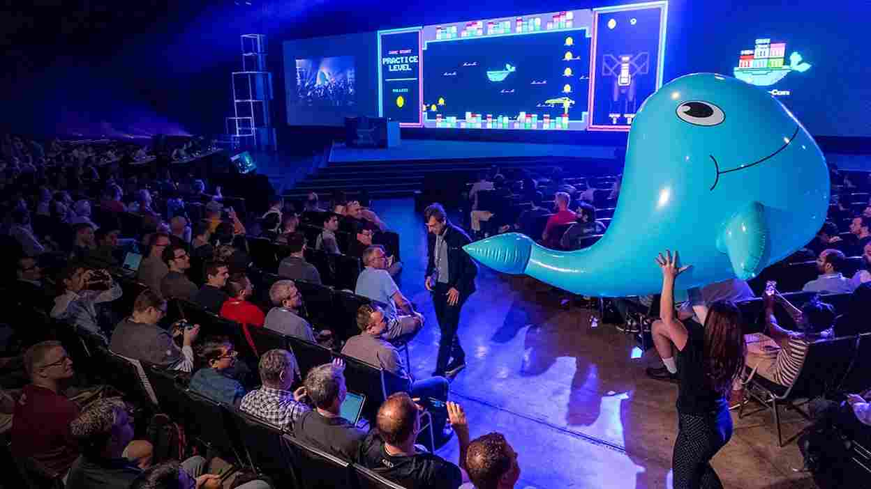 In a hall full of attendees at the DockerCon event an organizer holds up an inflatable whale and video-game imagery is shown on the screen.