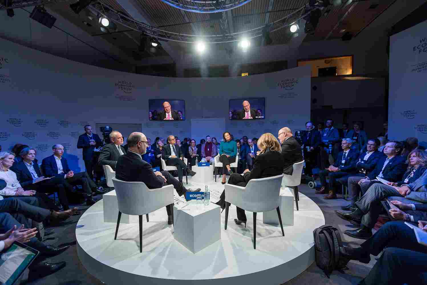A conference room with speakers sitting on a round stage while attendees sit in a circle around them at a live event.
