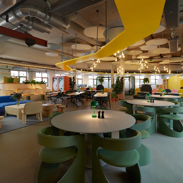 This is a photo of Toca Boca Campus. It is a wide open space with playful furniture and looks to be an exciting and playful place to eat lunch. There is a yellow wooden track on the ceiling and industrial pipes also. The colors are bright and playful, with some beige wooden furniture also.