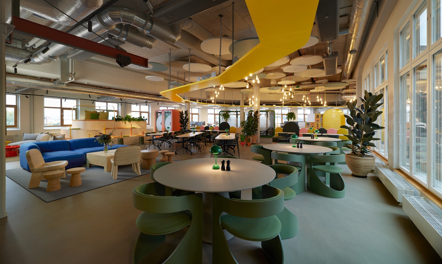This is a photo of Toca Boca Campus. It is a wide open space with playful furniture and looks to be an exciting and playful place to eat lunch. There is a yellow wooden track on the ceiling and industrial pipes also. The colors are bright and playful, with some beige wooden furniture also.