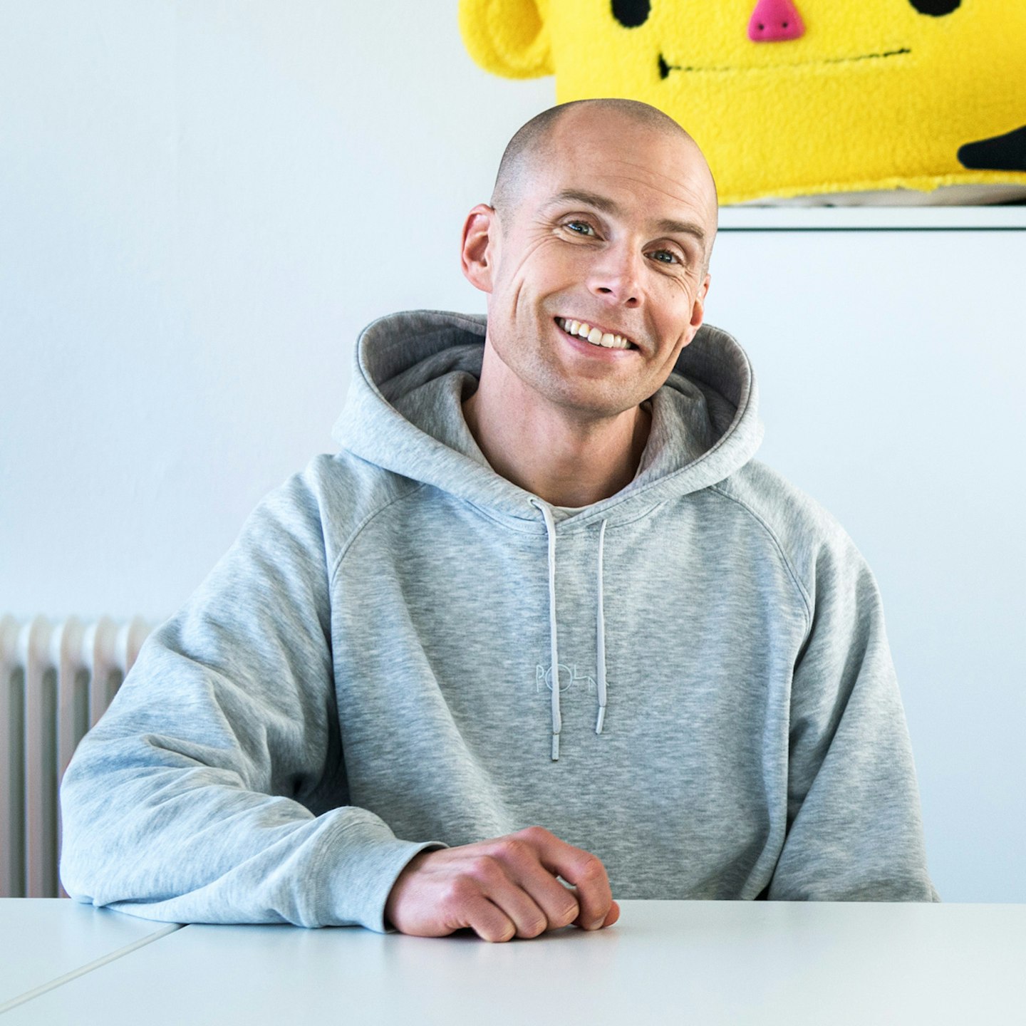 This is a photo of Emil Ovemar, Co-founder and Head of Studio for Toca Boca. He is sitting relaxed at a white table smiling wearing a grey hoodie. In the background there is a fluffy costume head of Bo, one of the characters from Toca Boca World