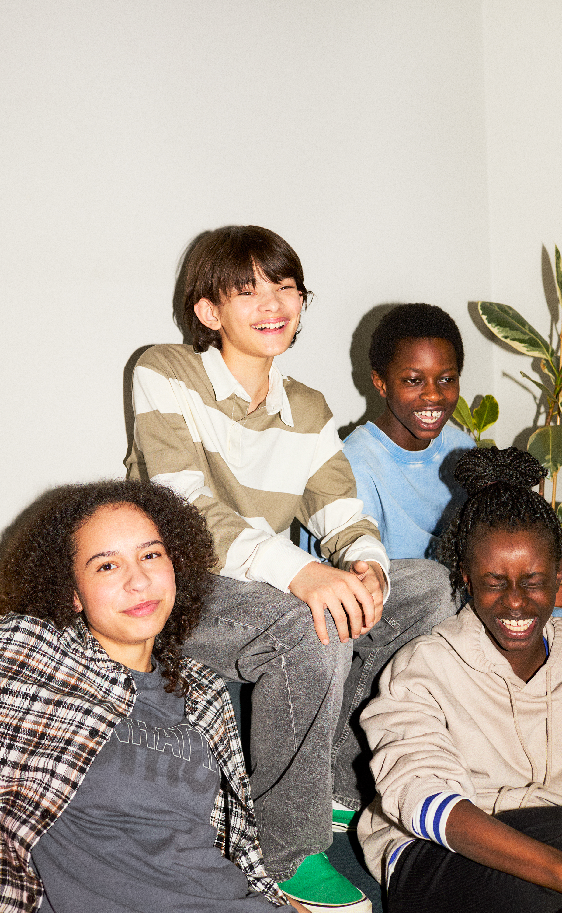 A group of diverse tween kids are hanging out on a sofa. Some are looking at the camera laughing while some are laughing and struggling to keep a straight face. One is even closing their eyes they are laughing so hard. They are wearing casual clothes with a cool relaxed street vibe