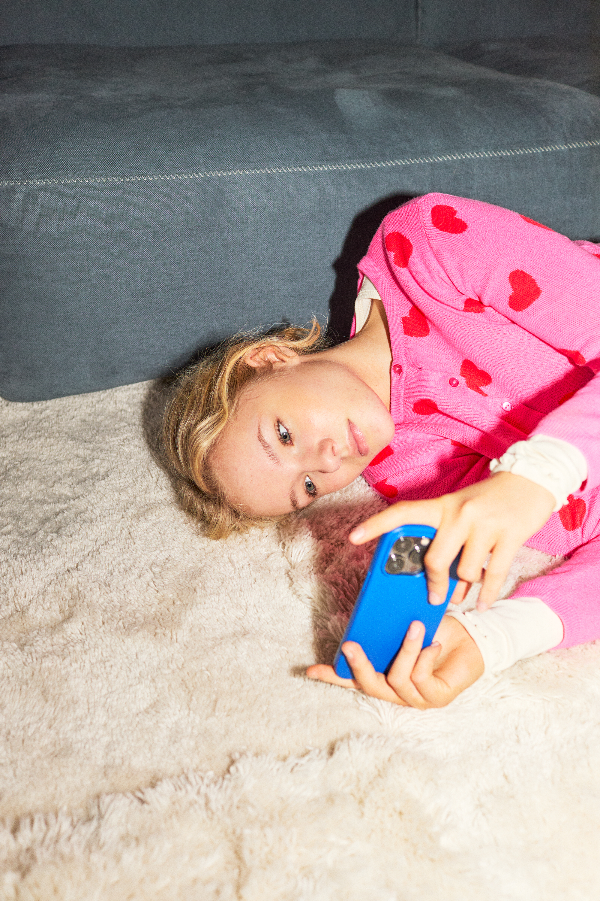 There is a photo of a blonde girl in her tween years playing on her phone laying down on a soft carpet. She is wearing a pink knitted jersey with hearts. The girl looks focused and the photo has bright highlights.