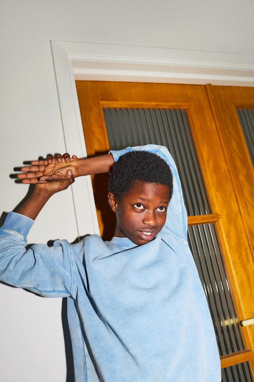 This is an image of a young tween kid, possibly a boy is stretching is arm over his head. He is wearing a light blue jumper. He has his chin down and appears to be listening to someone.
