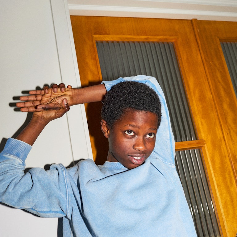 This is an image of a young tween kid, possibly a boy is stretching is arm over his head. He is wearing a light blue jumper. He has his chin down and appears to be listening to someone.