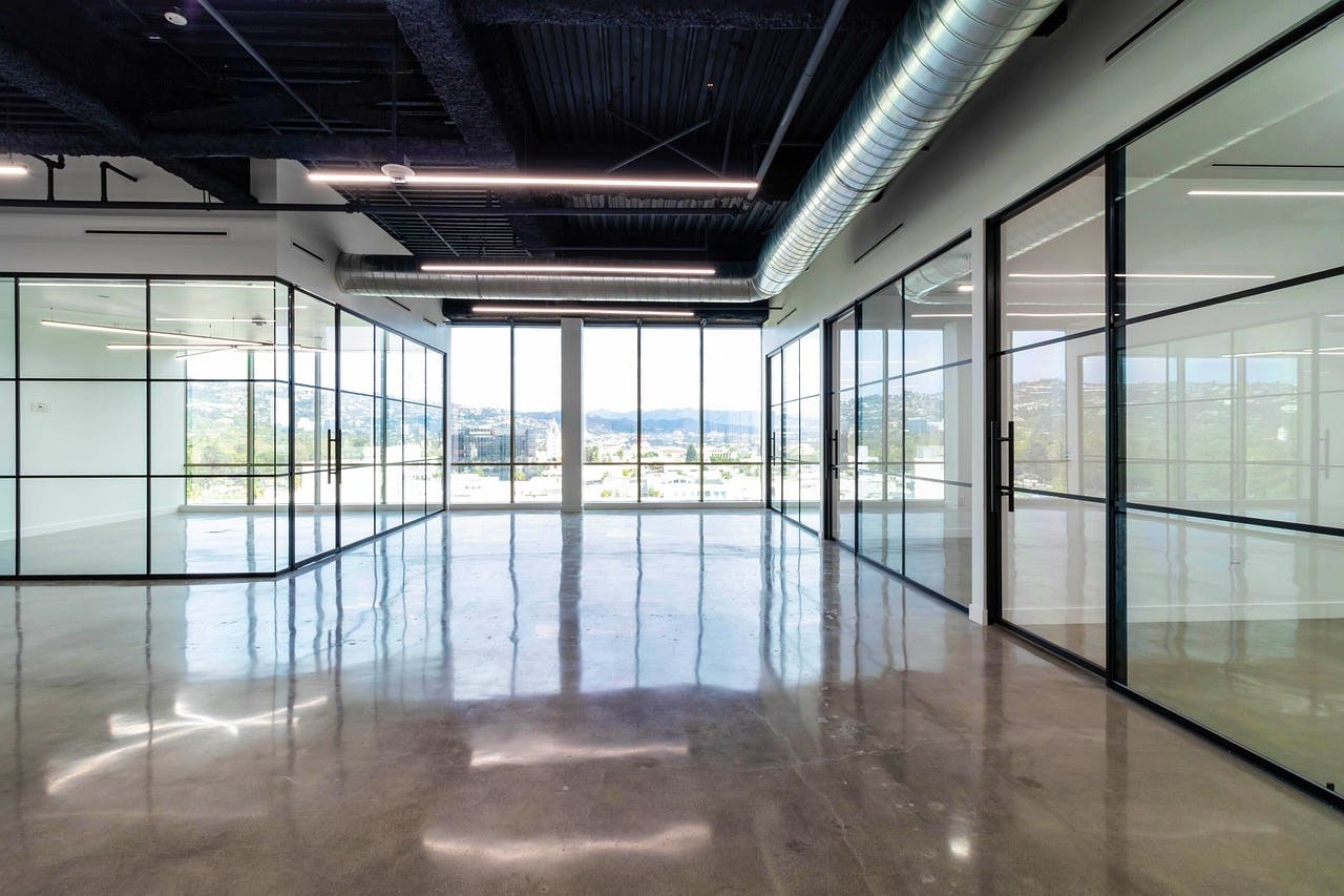 Modern industrial-style office interior featuring floor-to-ceiling glass walls, polished concrete floors, and large windows providing panoramic city and mountain views.