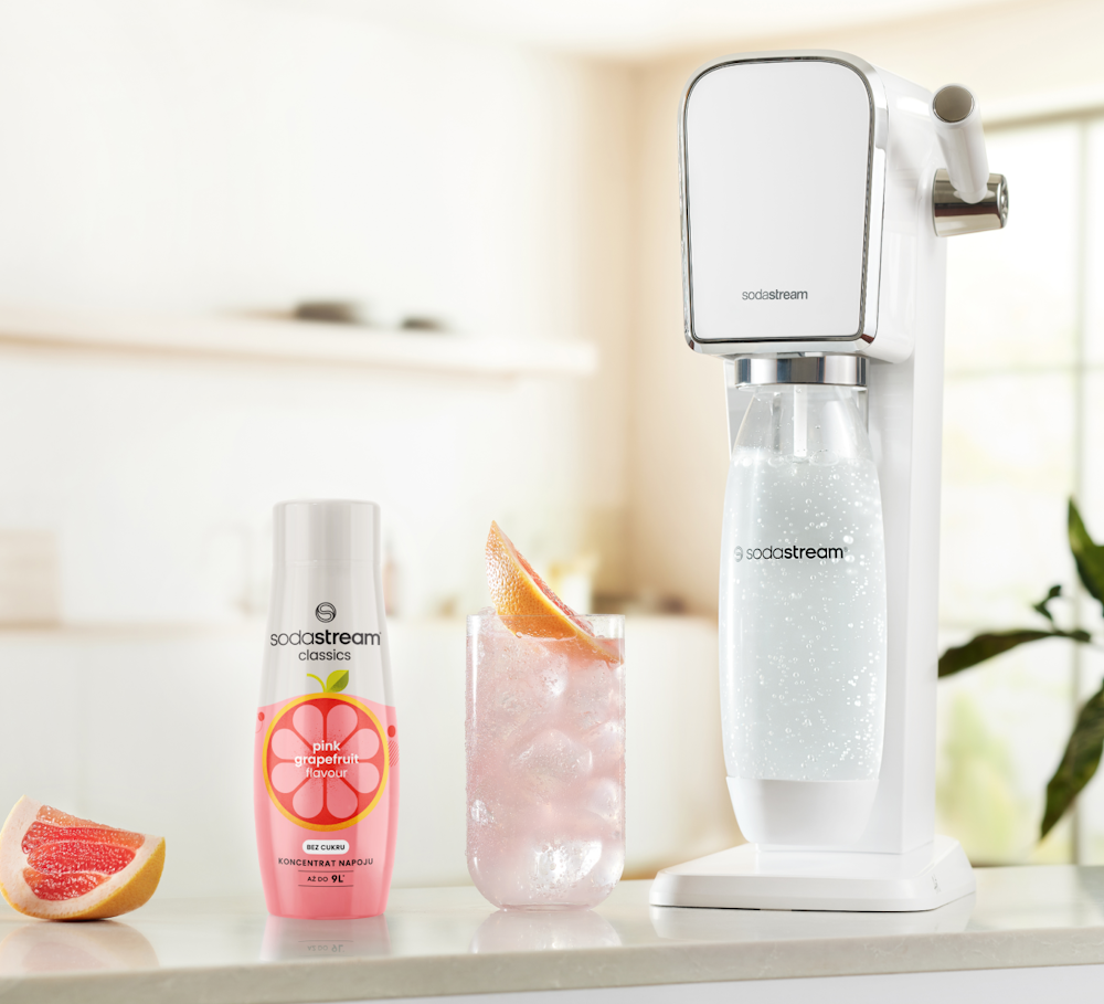 Koncentrat Różowy Grejpfrut wraz z saturatorem SodaStream i gotowym napojem w szklance stojące na kuchennym blacie.