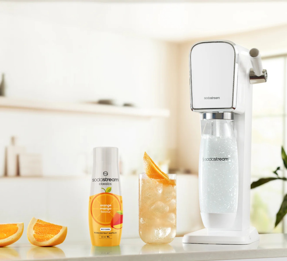 Koncentrat syropu pomarańczowego z mango wraz z saturatorem SodaStream i gotowym napojem w szklance stojące na kuchennym blacie.