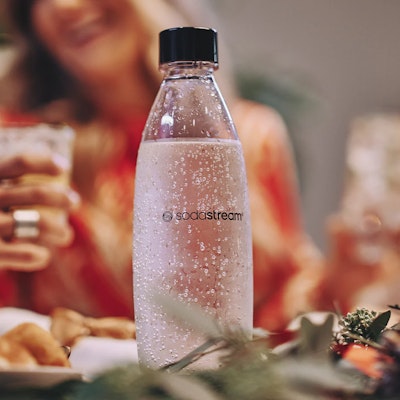Butelka wody gazowanej sodastream, w tle kobieta ze szklanką