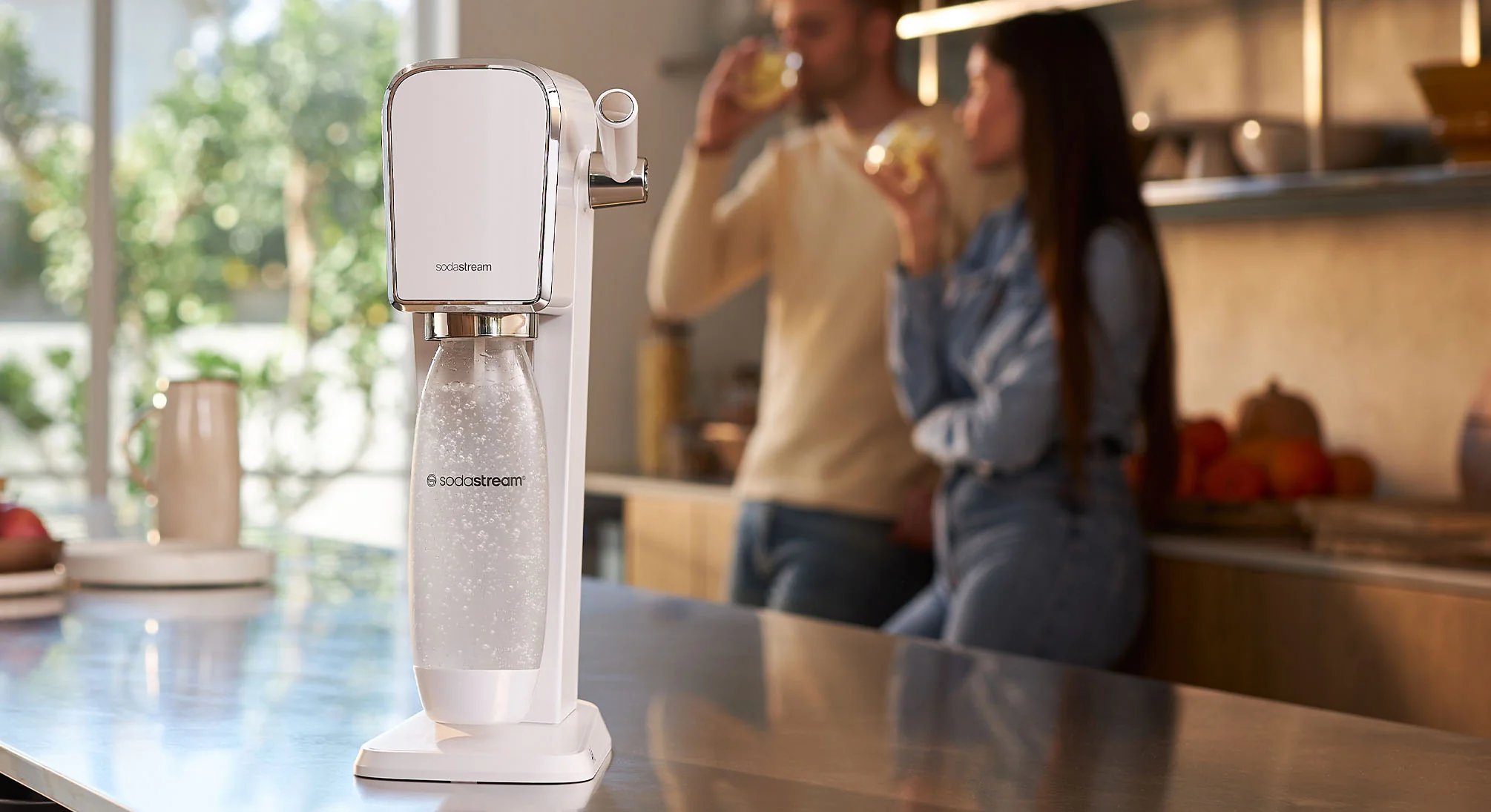 Stojący na ladzie saturator SodaStream Art, w tle para pijąca wodę gazowaną - tytułowa.