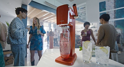 W tle grupa ludzi, a na pierwszym planie saturator do wody sodastream oraz dwa napoje z lodem i cytryną w przezroczystej szklance.