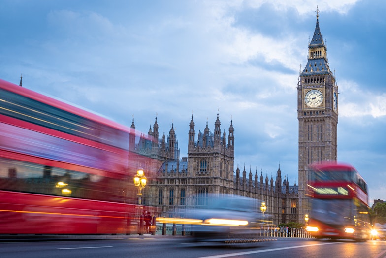 Red busses whirring past London's Big Ben at twilight