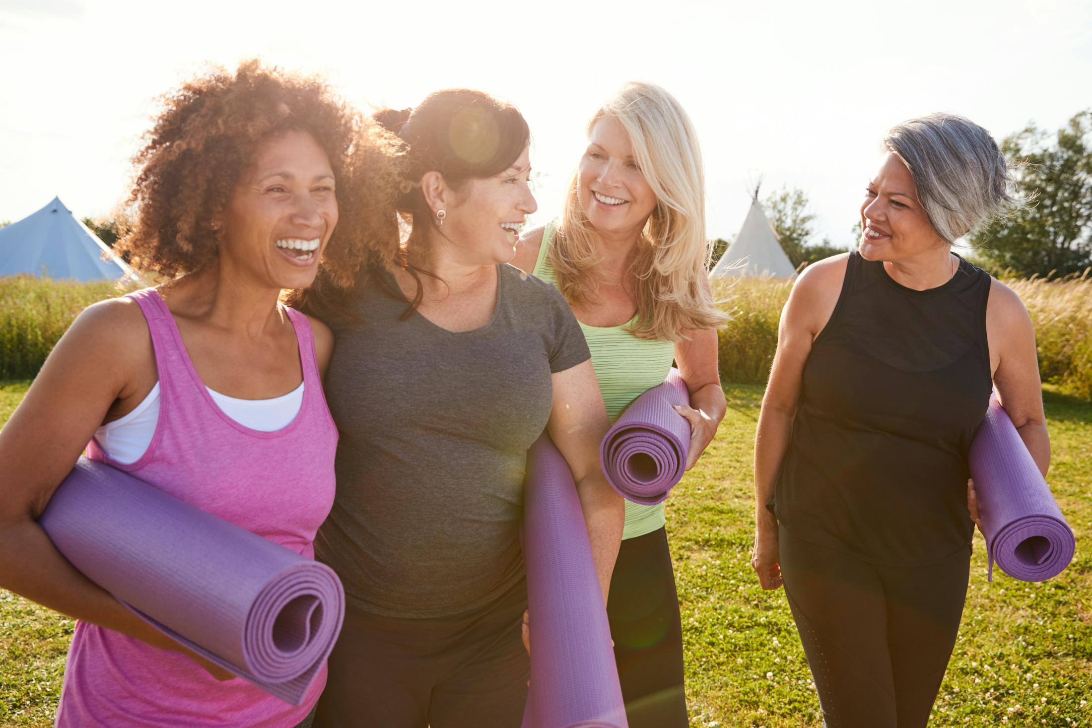 4 women in their 50's talking and laughing. They are in a grass field with yoga mats under their arms and workout clothes on.